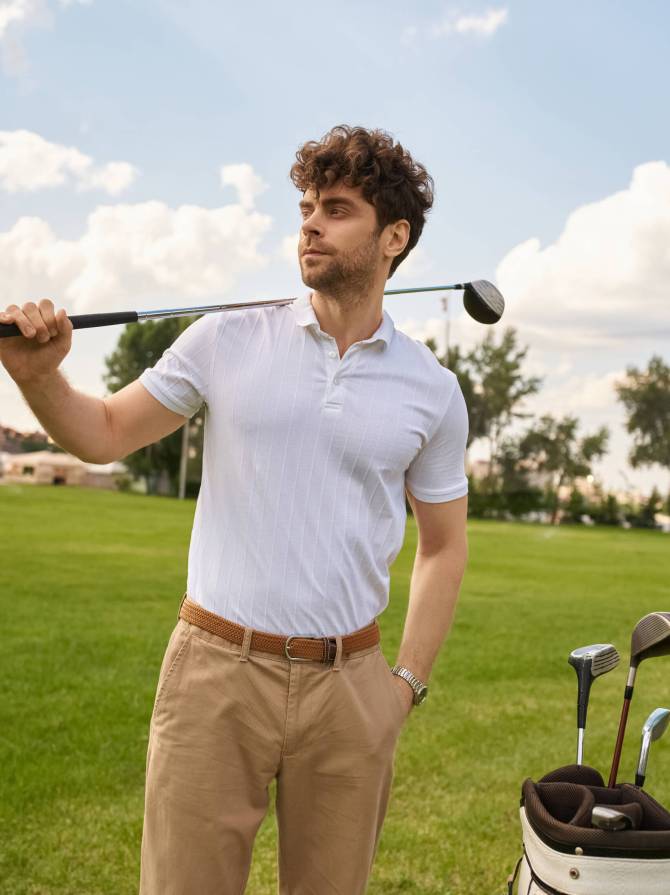 A man in elegant clothing holds a golf bag and club on a lush green field at a prestigious golf club.